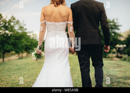 Hintere Ansicht von Braut und Bräutigam zu Fuß und Hand in Hand auf einer Wiese. Frisch vermählte Paar walking im Park am Tag ihrer Hochzeit. Stockfoto