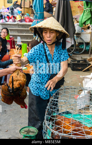 Eine Frau mit einer traditionellen Vietnamesischen konische bamboo Hut hält eine chcken durch die Beine, wie sie verkauft es an einen Kunden in Hoi An, Vietnam Stockfoto