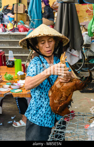 Eine Frau mit einer traditionellen Vietnamesischen konische bamboo Hut hält eine chcken durch die Beine, wie sie verkauft es an einen Kunden in Hoi An, Vietnam Stockfoto