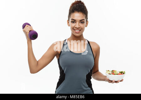 Gesunde und Fitness Concept - schöne sportliche African American auf Diät halten Hantel und frischen Salat auf Händen. Auf weissem studio Hintergrund isoliert. Stockfoto