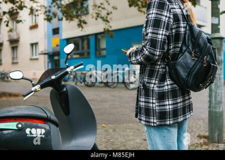 Ein Tourist mit einem Rucksack wird eine elektrische Scooter durch eine mobile Anwendung auf dem Telefon verwenden und distal zu aktivieren. Ein beliebtes Fahrzeug in Stockfoto