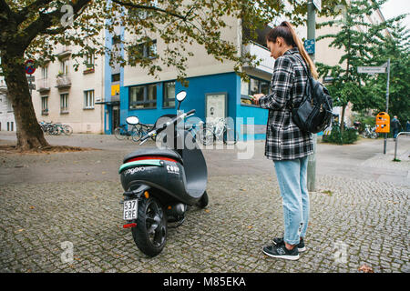 Ein Tourist mit einem Rucksack wird eine elektrische Scooter durch eine mobile Anwendung auf dem Telefon verwenden und distal zu aktivieren. Ein beliebtes Fahrzeug in Stockfoto