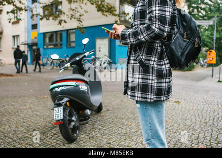 Ein Tourist mit einem Rucksack wird eine elektrische Scooter durch eine mobile Anwendung auf dem Telefon verwenden und distal zu aktivieren. Ein beliebtes Fahrzeug in Stockfoto