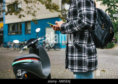 Ein Tourist mit einem Rucksack wird eine elektrische Scooter durch eine mobile Anwendung auf dem Telefon verwenden und distal zu aktivieren. Ein beliebtes Fahrzeug in Stockfoto