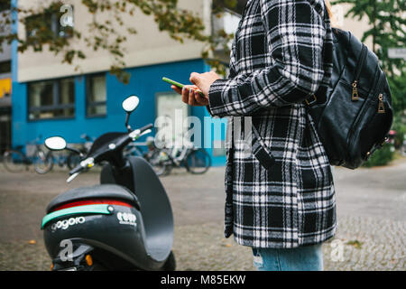 Ein Tourist mit einem Rucksack wird eine elektrische Scooter durch eine mobile Anwendung auf dem Telefon verwenden und distal zu aktivieren. Ein beliebtes Fahrzeug in Stockfoto