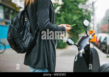 Ein Tourist mit einem Rucksack wird eine elektrische Scooter durch eine mobile Anwendung auf dem Telefon verwenden und distal zu aktivieren. Ein beliebtes Fahrzeug in Stockfoto
