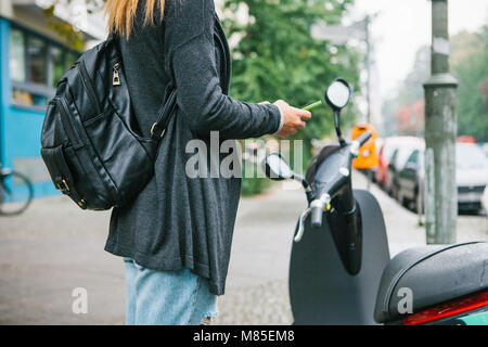 Ein Tourist mit einem Rucksack wird eine elektrische Scooter durch eine mobile Anwendung auf dem Telefon verwenden und distal zu aktivieren. Ein beliebtes Fahrzeug in Stockfoto