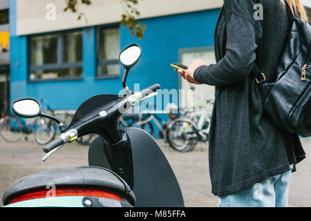 Ein Tourist mit einem Rucksack wird eine elektrische Scooter durch eine mobile Anwendung auf dem Telefon verwenden und distal zu aktivieren. Ein beliebtes Fahrzeug in Stockfoto