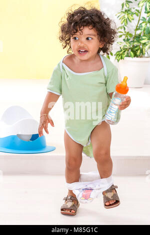 Junge stehend in Grün gestrickt Jumpsuit, diapper auf Beinen, halten Wasser aus einer Flasche iwitn einen weißen und blauen Töpfchen auf Hintergrund Stockfoto