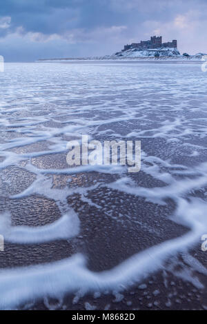 Schaum Muster auf Bamburgh Beach mit einem schneebedeckten Bamburgh Castle am Horizont, Bamburgh, Northumberland, England. Winter (März) 2018. Stockfoto
