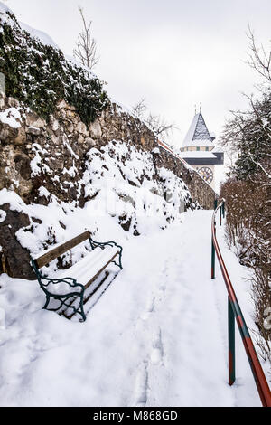 Verschneite Bank und Pfad zum Uhrturm Uhrturm Wahrzeichen der Stadt Graz auf Hill Schlossberg im Winter Stockfoto