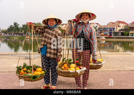 Zwei Obst Verkäufer tragen traditionelle Bambus konischen Hüten, Obst in bambuskörben von einem Pol in Hoi An, Vietnam. Es ist üblich, dass diese Frau Geld zu verlangen, nachdem sie ihre Fotografien und unvorsichtige Touristen einzuschüchtern. Stockfoto