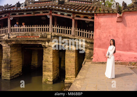 Eine sehr hübsche junge Dame in ein traditionelles, weißes Kleid steht neben Chùa Cầu japanische Brücke, eine hölzerne Brücke aus dem 18. Jahrhundert, der einen Schrein, in Hoi An, Vietnam geschnitzt Stockfoto