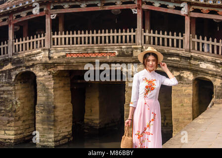 Eine sehr hübsche junge Dame in ein traditionelles, weißes Kleid steht neben Chùa Cầu japanische Brücke, eine hölzerne Brücke aus dem 18. Jahrhundert, der einen Schrein, in Hoi An, Vietnam geschnitzt Stockfoto