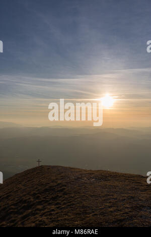 Kreuz auf dem Gipfel des Mt. Serrasanta (Umbrien, Italien), mit warmen goldenen Stunde Farben und die Sonne tief am Horizont Stockfoto