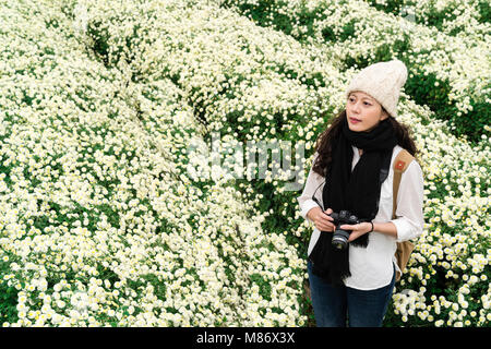 Portrait von schöne asiatische Frau mit Wolle Mütze und Schal mit den weißen Chrysantheme Garten im Herbst. Stehen und den Blick auf die Landschaft. Stockfoto