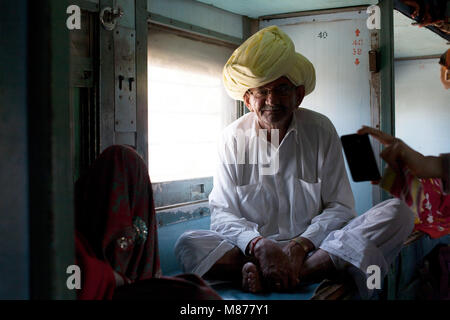 Eine alte indische Mann in Weiß einfache Kleidung und der Gelben Turbane, Brillen im Zug sitzt neben Fenster direkt auf die Kamera und lächelnd. Stockfoto