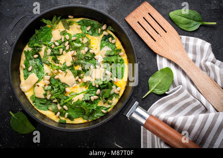 Spinat tortilla in der Pan auf dunklem Hintergrund. Omelette mit Eier, Spinat, Pinienkernen und Gemüse. Köstliches Frühstück Vorspeise oder Snack. Ansicht von oben Stockfoto