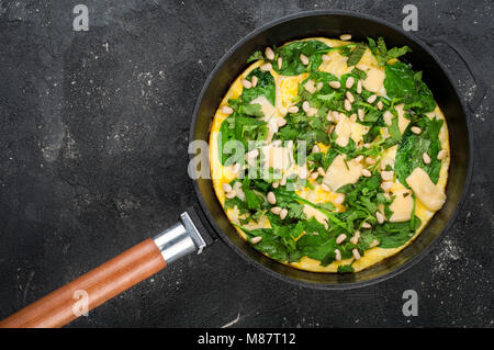 Omelette mit Eier, Spinat, Pinienkernen und Gemüse. Spinat tortilla in der Pan auf dunklem Hintergrund. Köstliches Frühstück Vorspeise oder Snack. Ansicht von oben Stockfoto