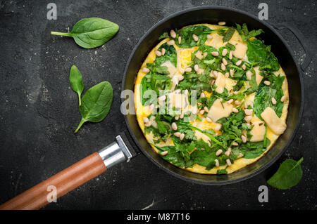 Omelette mit Eier, Spinat, Pinienkernen und Gemüse. Spinat tortilla in der Pan auf dunklem Hintergrund. Köstliches Frühstück Vorspeise oder Snack. Ansicht von oben Stockfoto