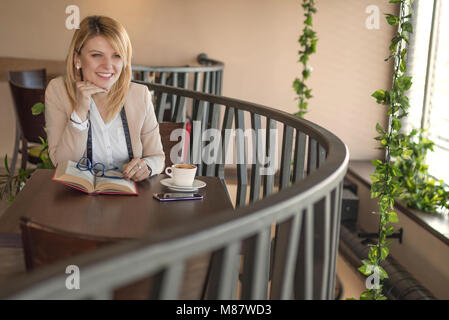 Junge lächelnde blonde Frau mit Brille in einem Restaurant ein Buch lesen und Kaffee trinken während einer Arbeitspause. Kaffeepause. Erholsame Zeit. Geschäft Stockfoto