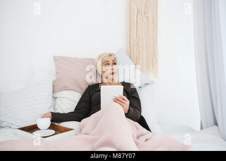 Eine ältere Frau im Bett lag, trinken Kaffee am Morgen eine Tablette zu Nachrichten oder Chatten mit Freunden in einem sozialen Netzwerk. Ältere generation und neue Stockfoto