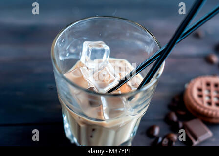 Kaffee mit Eis in ein Glas Milch, gegossen in ein Glas, Eiswürfel, Stücke von Schokolade und Plätzchen auf einem dunklen Holztisch Low Key Stockfoto