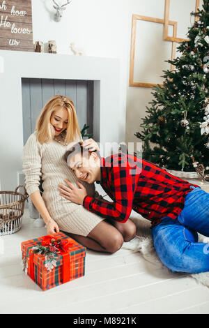 Glücklich Mann hört auf den Magen von seiner charmanten schwanger Breite beim Sitzen auf dem Teppich im liebevoll dekorierten Zimmer zu Weihnachten. Stockfoto