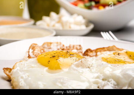 Traditionelles israelisches Frühstück mit zwei Spiegeleier, gelben Käse, Salat, eine frische Brötchen und einer Tasse Cappuccino. Nahaufnahme auf die gebratenen Eier Stockfoto