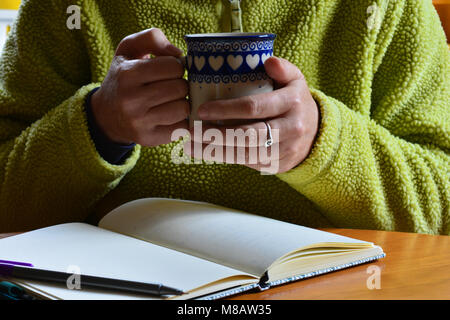 Frau sitzt an einem Tisch mit einer Tasse in der Hand und eine offene Notebook mit leeren Seiten vor ihr, im Innenbereich Stockfoto