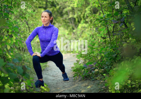 Schönen kaukasischen sportliche Mädchen im blauen Hemd und schwarzen Sport leggins führt Warming-up mit Beinen vor dem Joggen auf einem bunten grünen Wald zurück Stockfoto