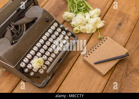 Schriftsteller flatlay. Dunkel Grau retro Schreibmaschine mit Notepad, schwarzer Stift und weißen Ranunkeln über Holz- Hintergrund. Stockfoto