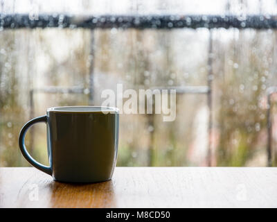Eine Tasse auf einem hölzernen Tisch vor einem Glas Fenster mit Tropfen bedeckt. Stockfoto