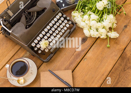 Schriftsteller flatlay. Dunkel Grau retro Schreibmaschine mit Notepad, schwarze Kugelschreiber, Tasse schwarzen Kaffee, vintage Film Kamera und weißen Ranunkeln über Holz- backgrou Stockfoto