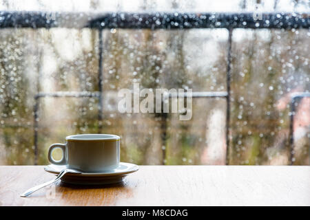 Eine weiße Espressotasse mit einem Löffel auf einem Holztisch vor einem Glasfenster, das mit Regentropfen bedeckt ist und eine geringe Schärfentiefe hat. Stockfoto