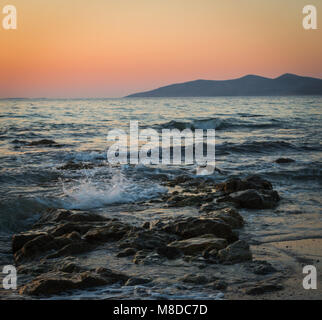 Eine lange Einwirkung auf das Meer wie es bricht über die Felsen und Strand in goldenen Stunde, und die Sonne hinter den Bergen. Stockfoto