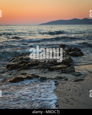 Eine lange Einwirkung auf das Meer wie es bricht über die Felsen und Strand in goldenen Stunde, und die Sonne hinter den Bergen. Stockfoto