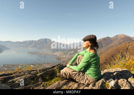 Frau mit flacher Deckel sitzen und genießen Sie den Panoramablick über die Berge und den Lago Maggiore im Tessin, Schweiz Stockfoto