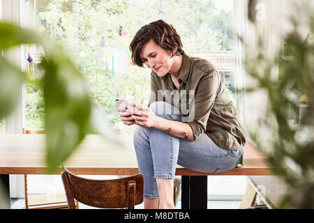 Mitte der erwachsenen Frau zu Hause, sitzt am Tisch, mit Smartphone Stockfoto