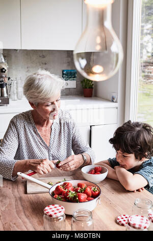 Großmutter am Küchentisch sitzen, die Vorbereitung von Erdbeeren, Enkel neben ihr sitzen, beobachten Stockfoto