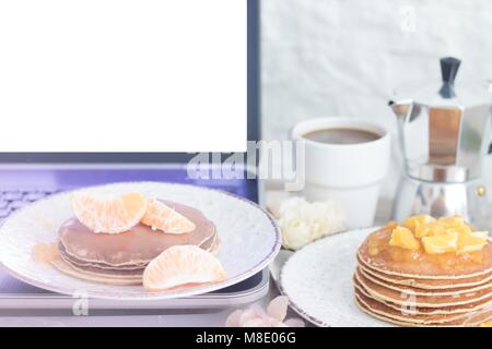 Laptop mit weißen leeren Bildschirm auf Tabelle mit Frühstück: Pfannkuchen mit in Scheiben geschnittenen Mandarinen und Orangen Marmelade und Kaffee mit Metall Coffee Pot. Vorderansicht. C Stockfoto