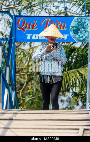 Eine Frau mit einer traditionellen vietnamesischen Bambus konische hat überprüft Ihr Smartphone, als sie über die Brücke geht, Vietnam Stockfoto