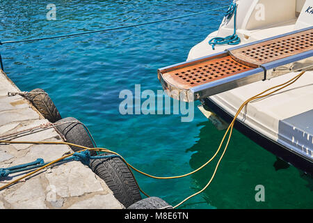 Boardingleiter aus weiß glänzendem Motorboot des modernen Designs, der mit Seilen auf blauem Meerwasser an der Pier festgemacht ist, das am Sommertag helles Sonnenlicht reflektiert Stockfoto