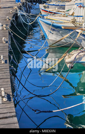 Verschiedene Motorboote, die auf braunem hölzernen Pier festgemacht sind, mit Seilen zu Küstenbissen auf Meerwasser, die im Sommer Sonnenlicht reflektieren Stockfoto