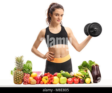 Fitness Frau mit einer Hantel hinter einem Tisch mit Obst und Gemüse auf weißem Hintergrund Stockfoto