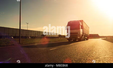 Weiß Semi Truck mit Cargo Trailer angeschlossene Laufwerke auf die leere Straße. Industriehallen an den Seiten der Autobahn. Sonnenuntergang. Stockfoto