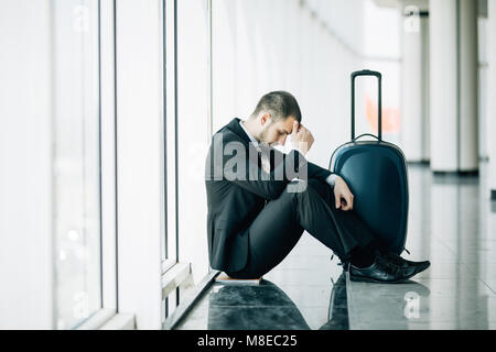 Business Mann am Terminal Flughafen auf dem Boden sitzend mit Koffer Flug Verspätung, zwei Hände am Kopf berühren, Kopfschmerzen, Warten auf Reisen. Stockfoto