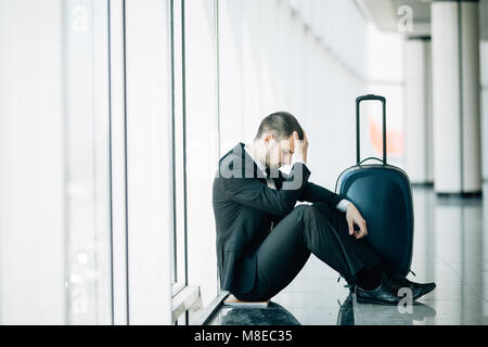 Business Mann am Terminal Flughafen auf dem Boden sitzend mit Koffer Flug Verspätung, zwei Hände am Kopf berühren, Kopfschmerzen, Warten auf Reisen. Stockfoto