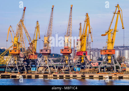 Gelben Krane, die in einem Seehafen vor dem Hintergrund von Metall Getreidespeicher und ein blauer Himmel mit Wolken und fliegen Scharen von Vögeln Stockfoto