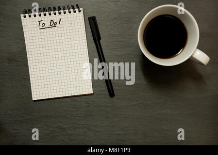Aufgabenliste in vermaschten Spirale Notizblock und Stift mit Tasse Kaffee, auf grauem Schiefer Table Top - von oben, minimale Stockfoto